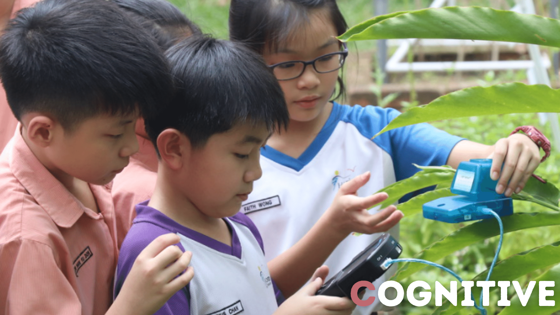 Three children in school uniforms examine a small blue sensor device attached to a large green plant leaf outdoors.